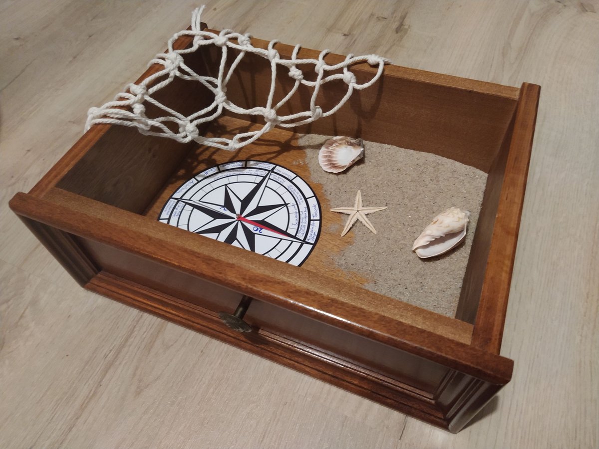 Eine Holzkiste mit Sand, einer Kompassrose, Muscheln, einem Seestern und einem weißen Fischernetz, maritimes Thema darstellend.