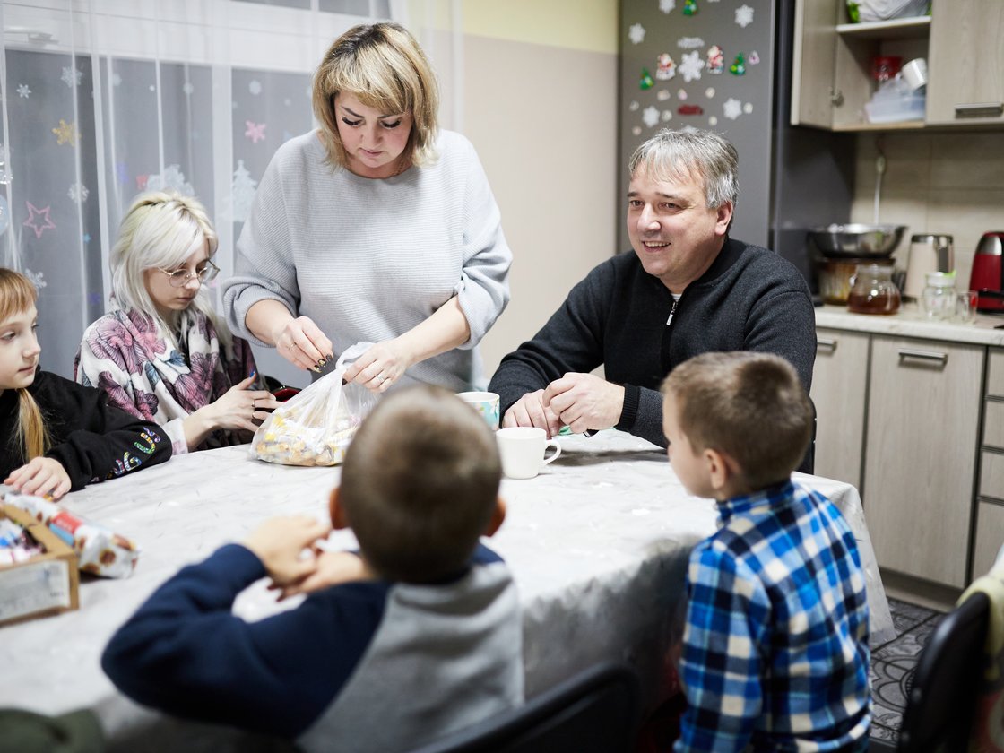 Sechsköpfige Familie am Tisch; Frau öffnet Snacktüte, Mann lächelt, Kinder schauen in häuslicher Küche.