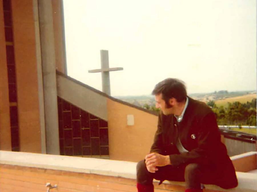 Mann mit Bart in Jacke und roten Stiefeln sitzt auf Mauer, blickt auf Landschaft mit moderner Kirche und Kreuz.