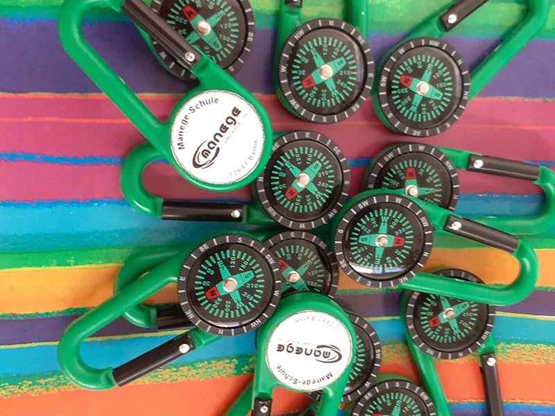 Eine Ansammlung grüner Karabiner-Kompass-Anhänger mit "Manege-Schule"-Logo auf einem bunten Streifenmuster.