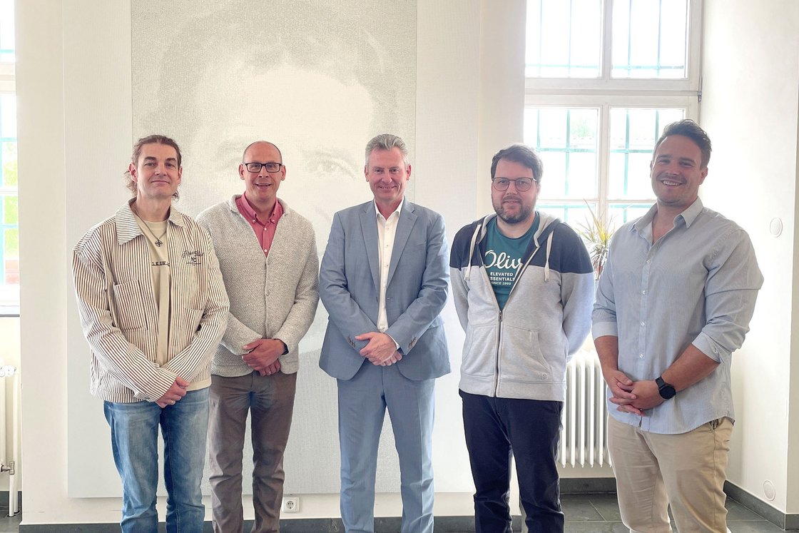 Fünf Männer stehen lächelnd in einem hellen Raum mit großen Fenstern und einem dezenten Porträt an der Wand.