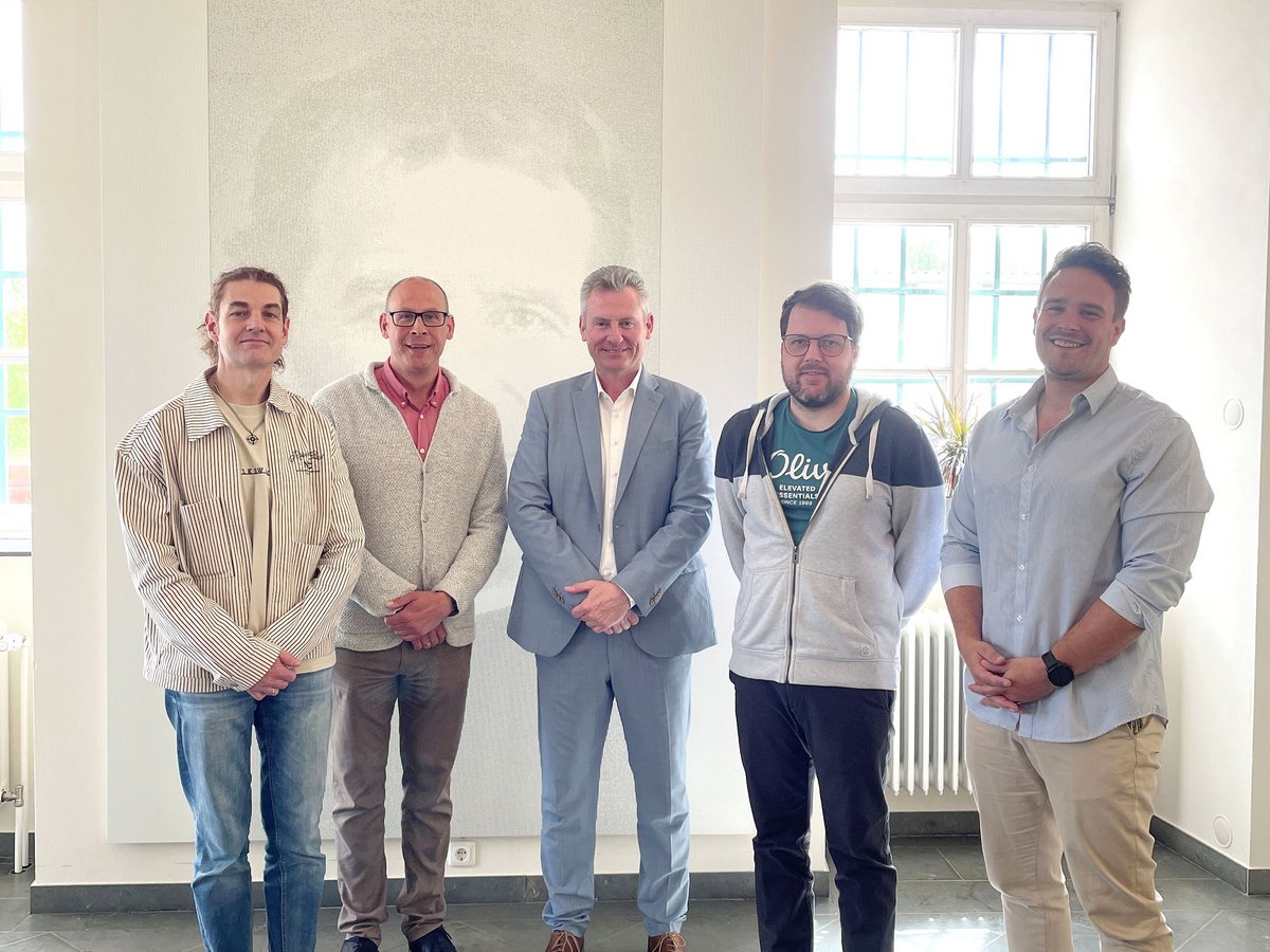 Fünf Männer stehen lächelnd in einem hellen Raum mit großen Fenstern und einem dezenten Porträt an der Wand.