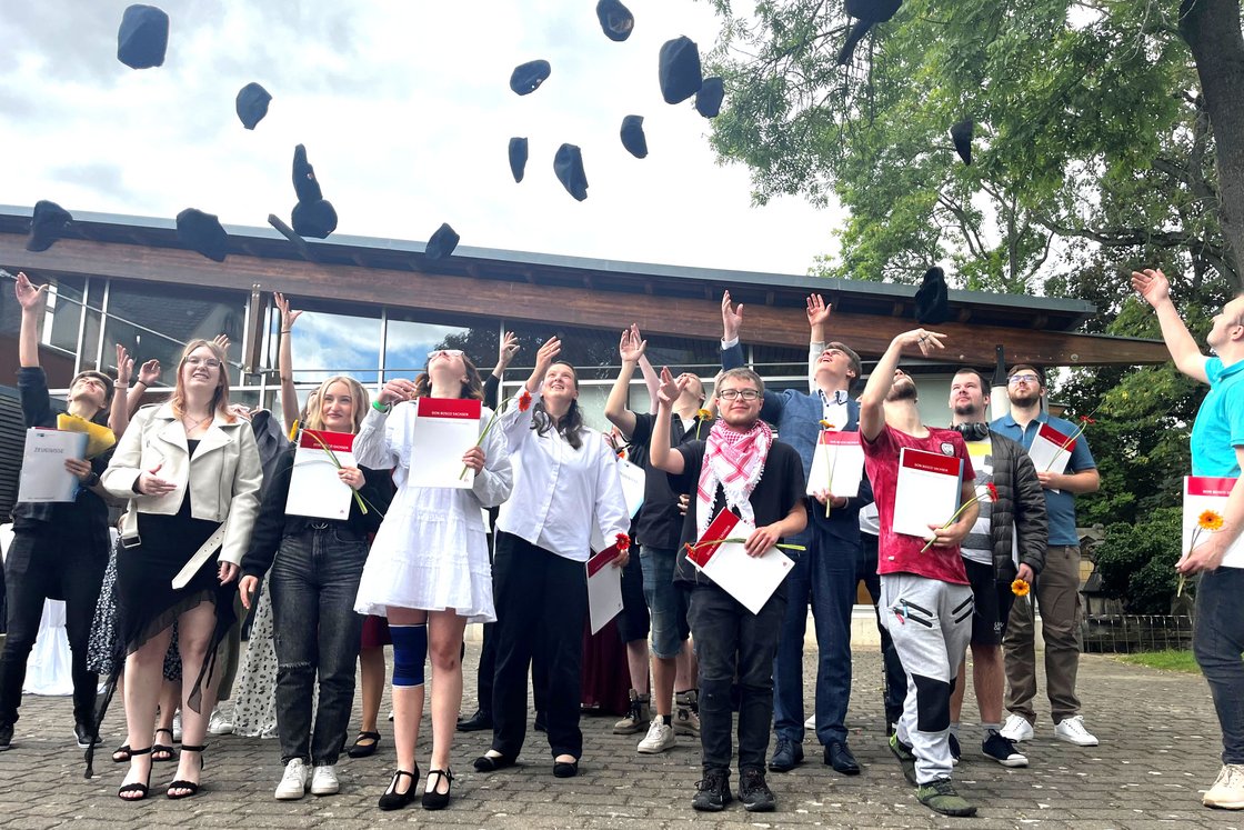 Glückliche Absolventen werfen ihre Hüte in die Luft, halten Zeugnisse und Blumen und feiern ihren erfolgreichen Abschluss im Freien.