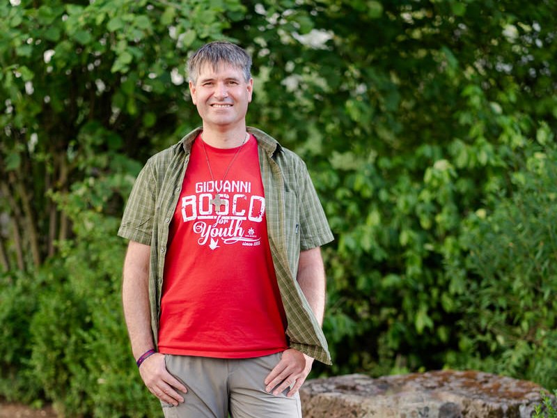 Ein lächelnder Mann in einem roten "Giovanni Bosco for Youth"-T-Shirt und offenem Karohemd steht vor grünem Laub.