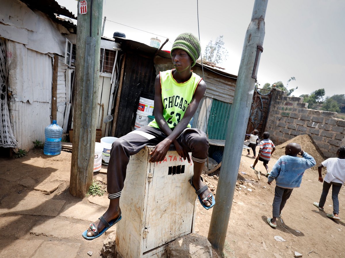 Ein Jugendlicher mit grüner Mütze und Basketball-Trikot sitzt auf einer Kiste vor Hütten, Kinder spielen.