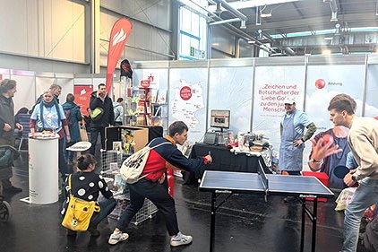 Eine lebhafte Messehalle mit Ständen und Besuchern, im Vordergrund spielen zwei Männer Tischtennis.