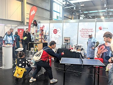 Eine lebhafte Messehalle mit Ständen und Besuchern, im Vordergrund spielen zwei Männer Tischtennis.