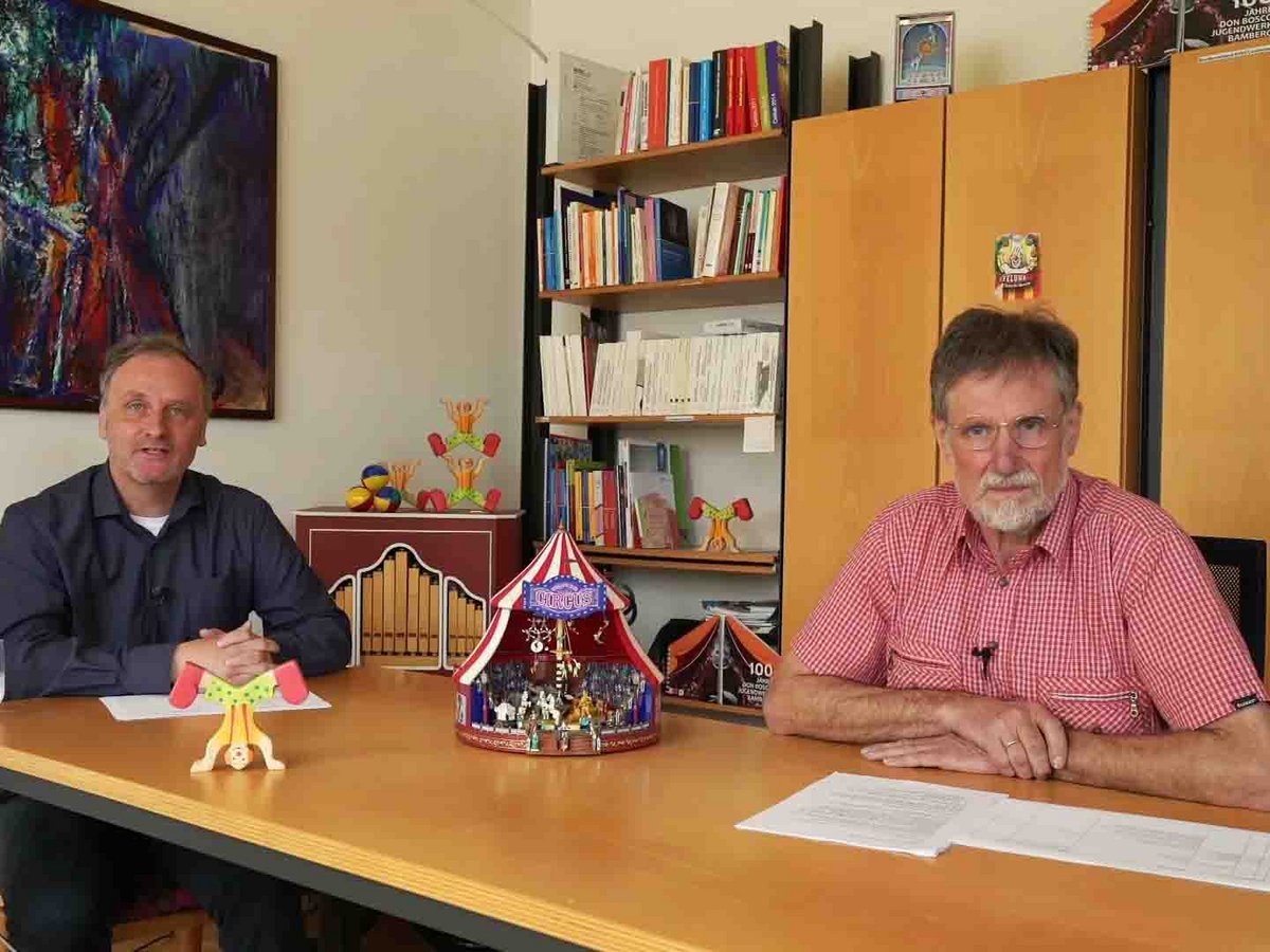 Zwei Männer sitzen an einem Tisch in einem mit Zirkusartikeln dekorierten Büro, einer spricht, der andere hört zu.