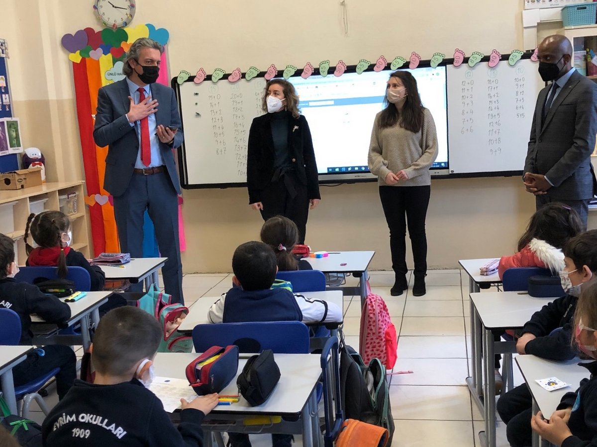 Vier maskierte Erwachsene interagieren mit jungen Schülern in einem Klassenzimmer. Ein Mann gestikuliert, während drei andere bei einer interaktiven Tafel stehen.