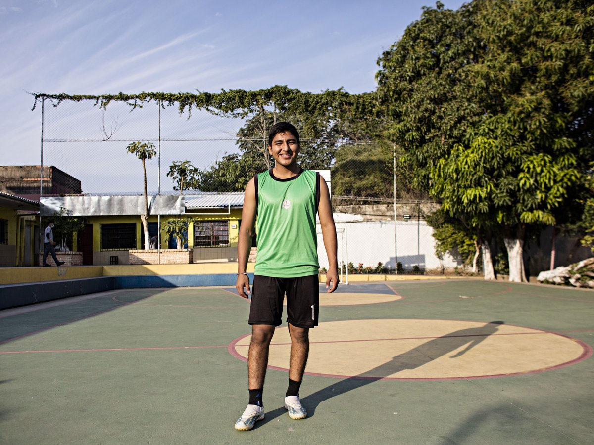 Lächelnder junger Mann in grünem Sporttop und schwarzen Shorts steht auf einem Mehrzweck-Sportplatz im Freien.