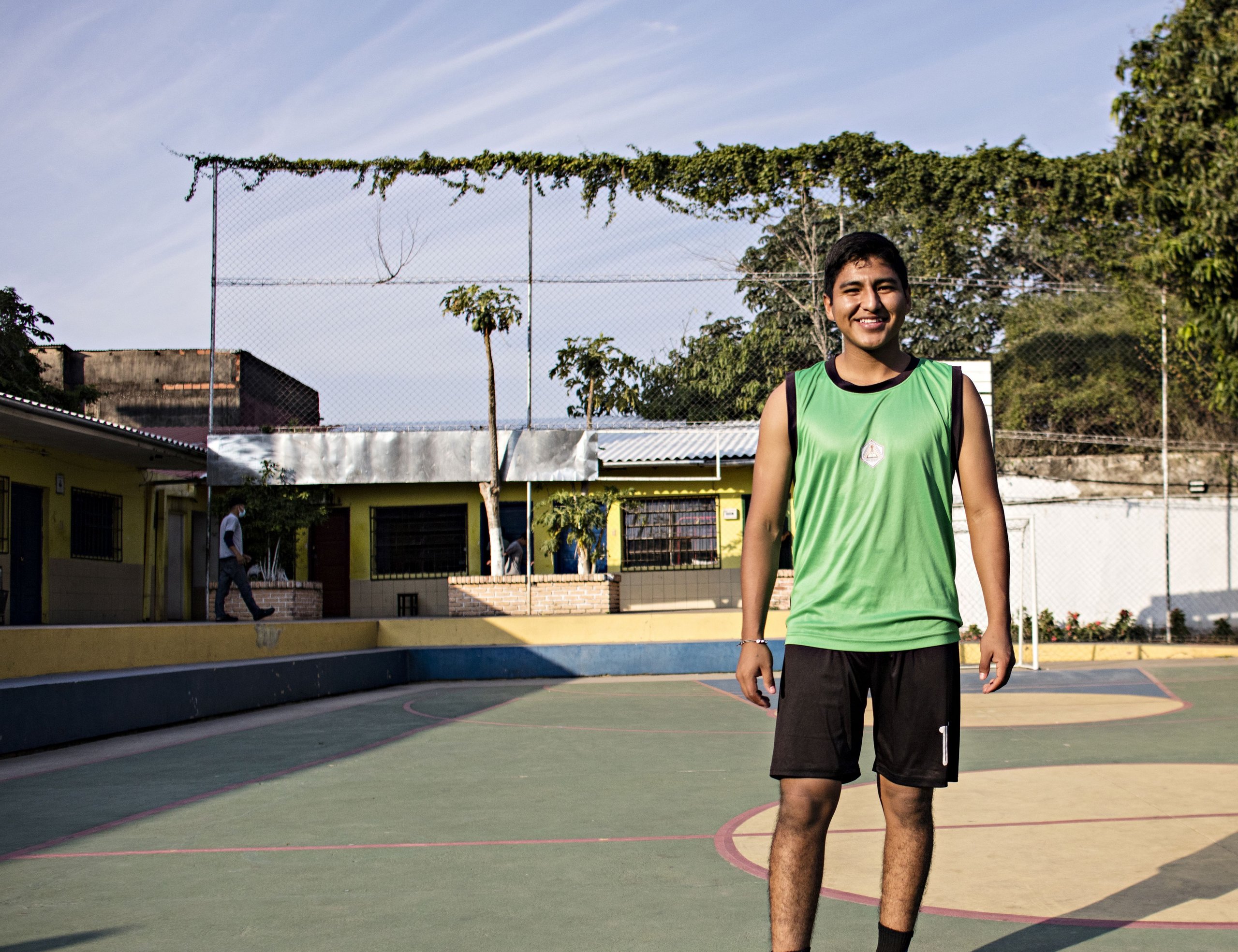 Lächelnder junger Mann in grünem Sporttop und schwarzen Shorts steht auf einem Mehrzweck-Sportplatz im Freien.