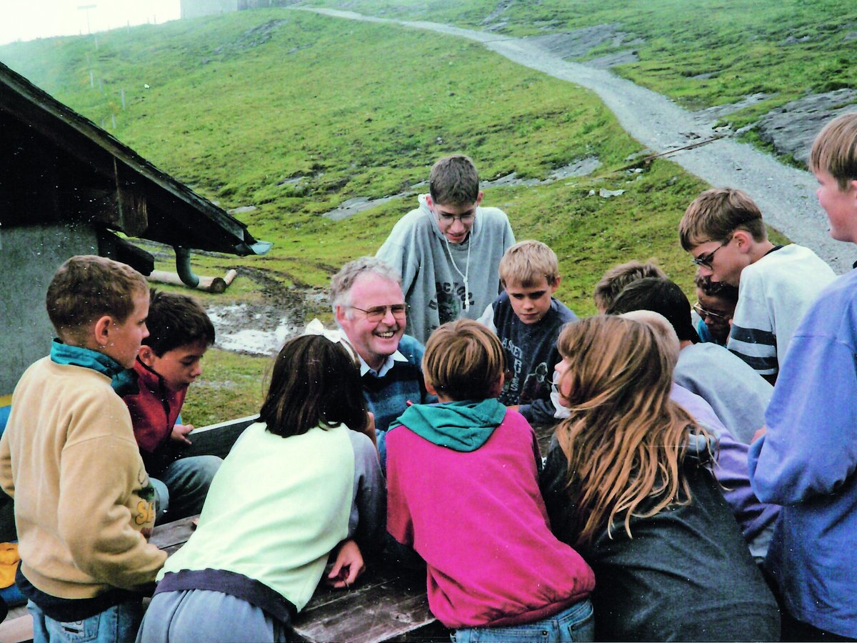 Ein lächelnder Mann sitzt an einem Holztisch, umgeben von Kindern und Jugendlichen, auf einer Wiese mit Pfad.