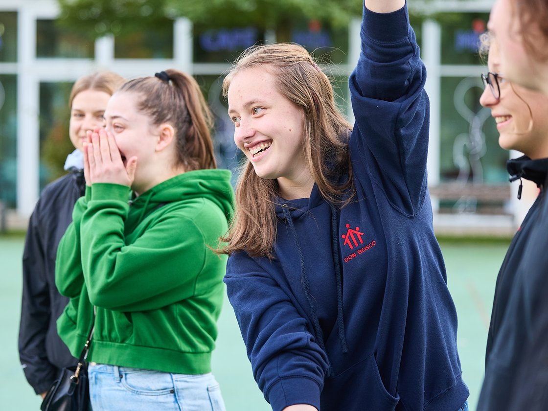 Vier junge Leute lachen draußen; eine im blauen Kapuzenpullover hebt den Arm, eine andere im grünen bedeckt den Mund.