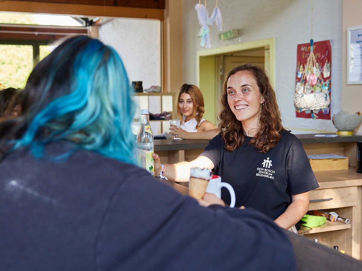 Eine lächelnde Frau im Don Bosco Zentrum Regensburg reicht einer Person mit blauen Haaren Getränke und Eis über die Theke.