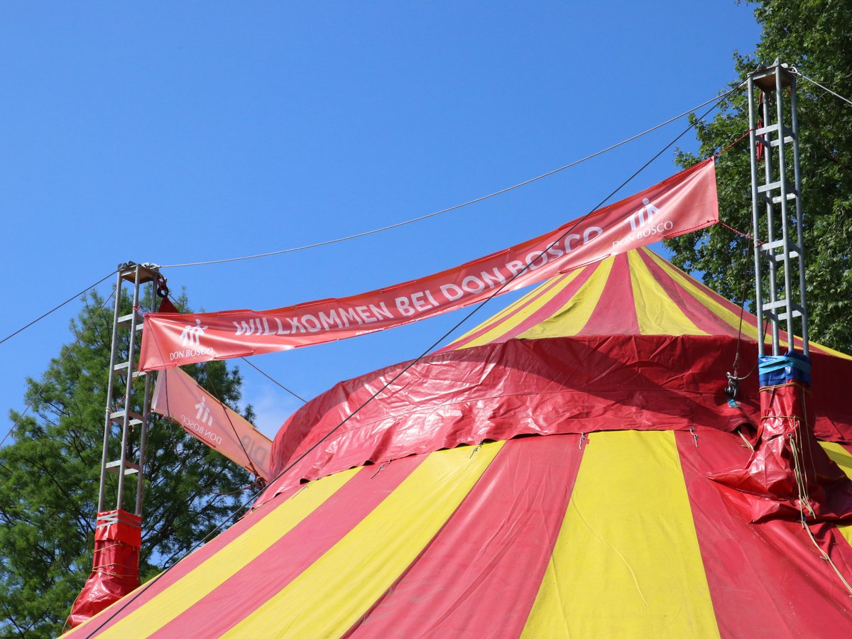Rot-gelbes Zirkuszelt mit rotem Banner 'WILLKOMMEN BEI DON BOSCO' vor blauem Himmel und Bäumen.