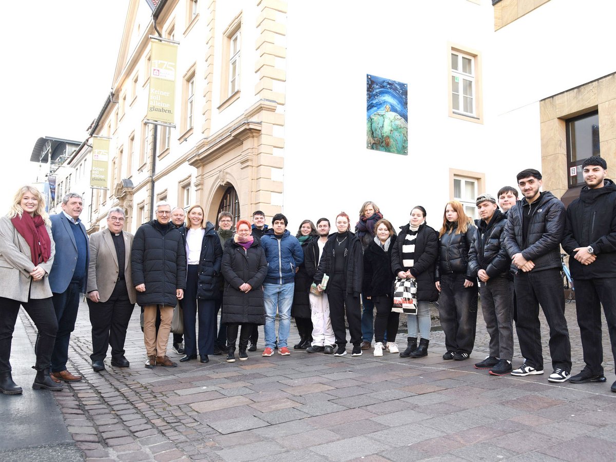 Eine vielfältige Gruppe von Erwachsenen und Jugendlichen steht auf einem gepflasterten Straße vor einem Gebäude mit Bannern.