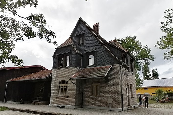 Historisches Backsteingebäude mit steilem Schieferdach und Gaubenfenstern, daneben ein Holzschuppen, unter bewölktem Himmel.