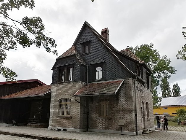 Historisches Backsteingebäude mit steilem Schieferdach und Gaubenfenstern, daneben ein Holzschuppen, unter bewölktem Himmel.