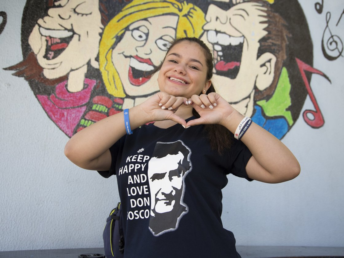Lächelndes Mädchen formt mit Händen ein Herz, trägt Don Bosco T-Shirt, vor einem Wandbild.
