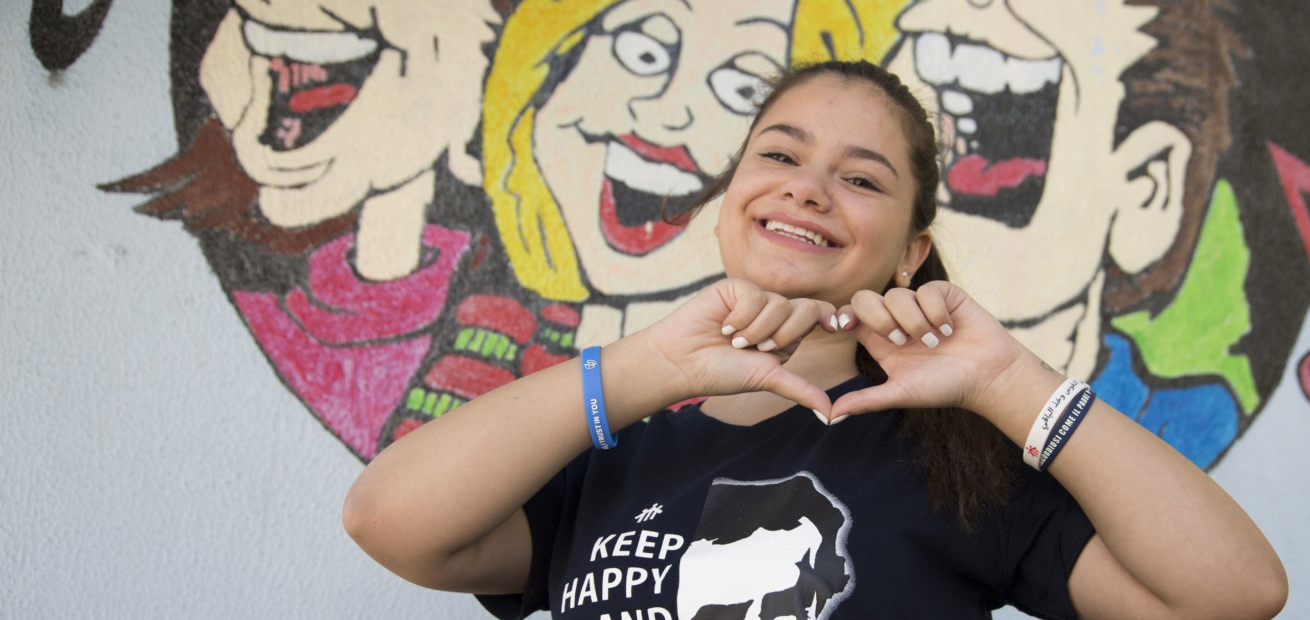 Lächelndes Mädchen formt mit Händen ein Herz, trägt Don Bosco T-Shirt, vor einem Wandbild.