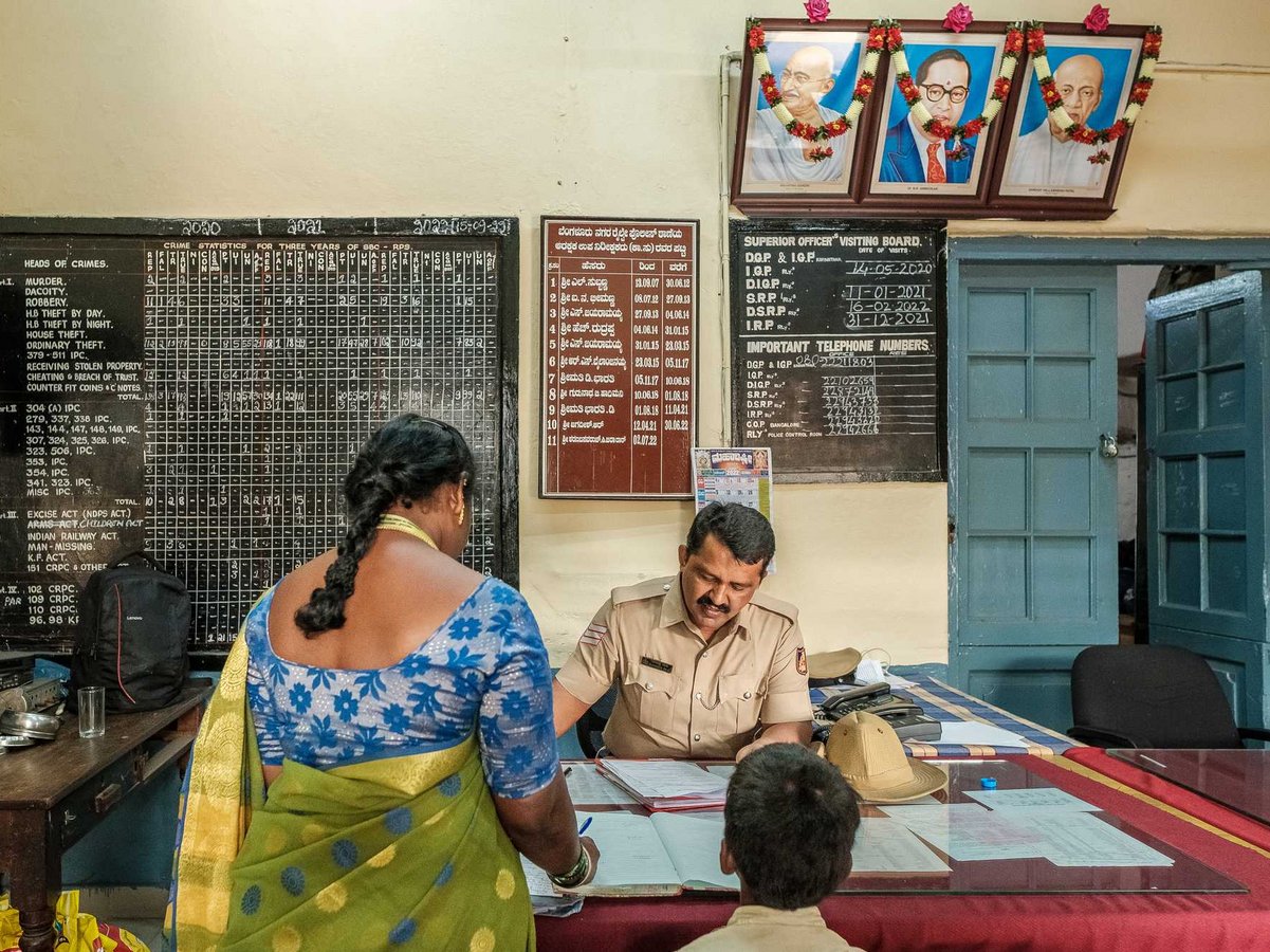 Ein indischer Polizist am Schreibtisch, der Dokumente mit einer Frau im Sari und einem Kind in einem geschäftigen Büro überprüft.