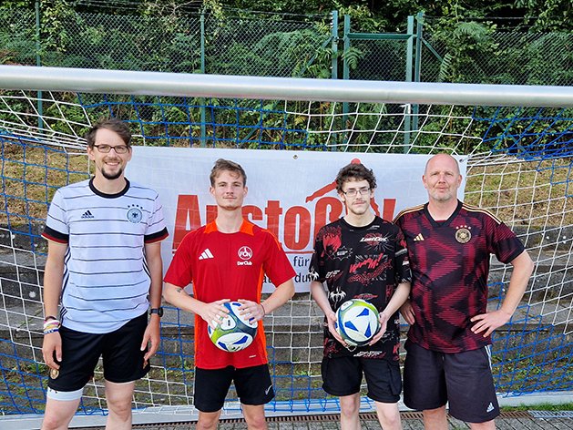 Vier Männer in Fußballtrikots stehen vor einem Tor, zwei halten Bälle. Ein Banner ist im Hintergrund.