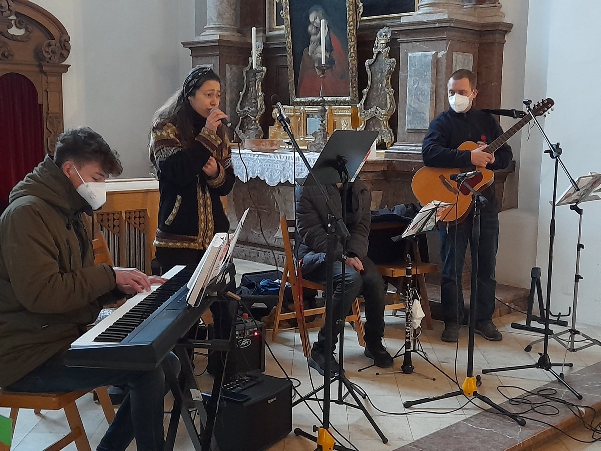 Eine Musikgruppe tritt in einer Kirche auf. Eine Frau singt ins Mikrofon, ein Mann spielt Keyboard, ein anderer Gitarre, alle tragen Masken.