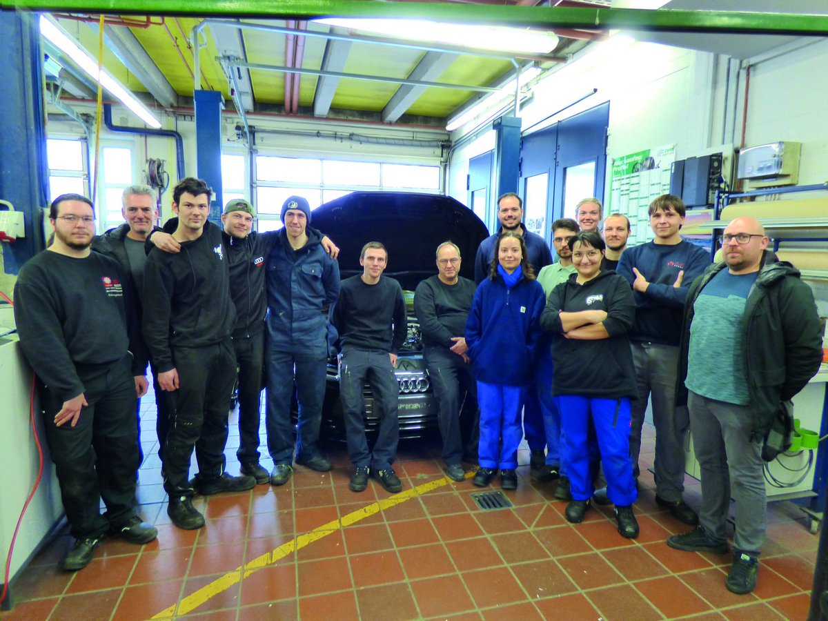 Eine Gruppe von fünfzehn Personen in einer Autowerkstatt, die vor einem Auto mit offener Motorhaube posiert.