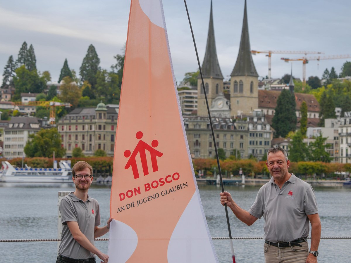 Zwei Männer halten eine große Don Bosco Flagge an einem Seeufersteg, mit Stadt und Kirchtürmen im Hintergrund.
