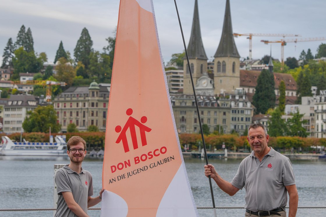 Zwei Männer halten eine große Don Bosco Flagge an einem Seeufersteg, mit Stadt und Kirchtürmen im Hintergrund.