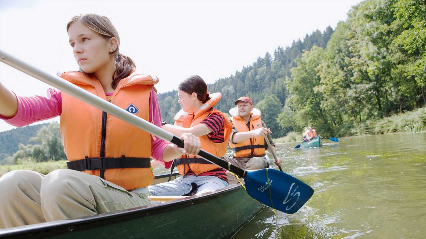 Personen in orangefarbenen Schwimmwesten paddeln in Kanus auf einem Fluss, umgeben von bewaldeten Ufern.