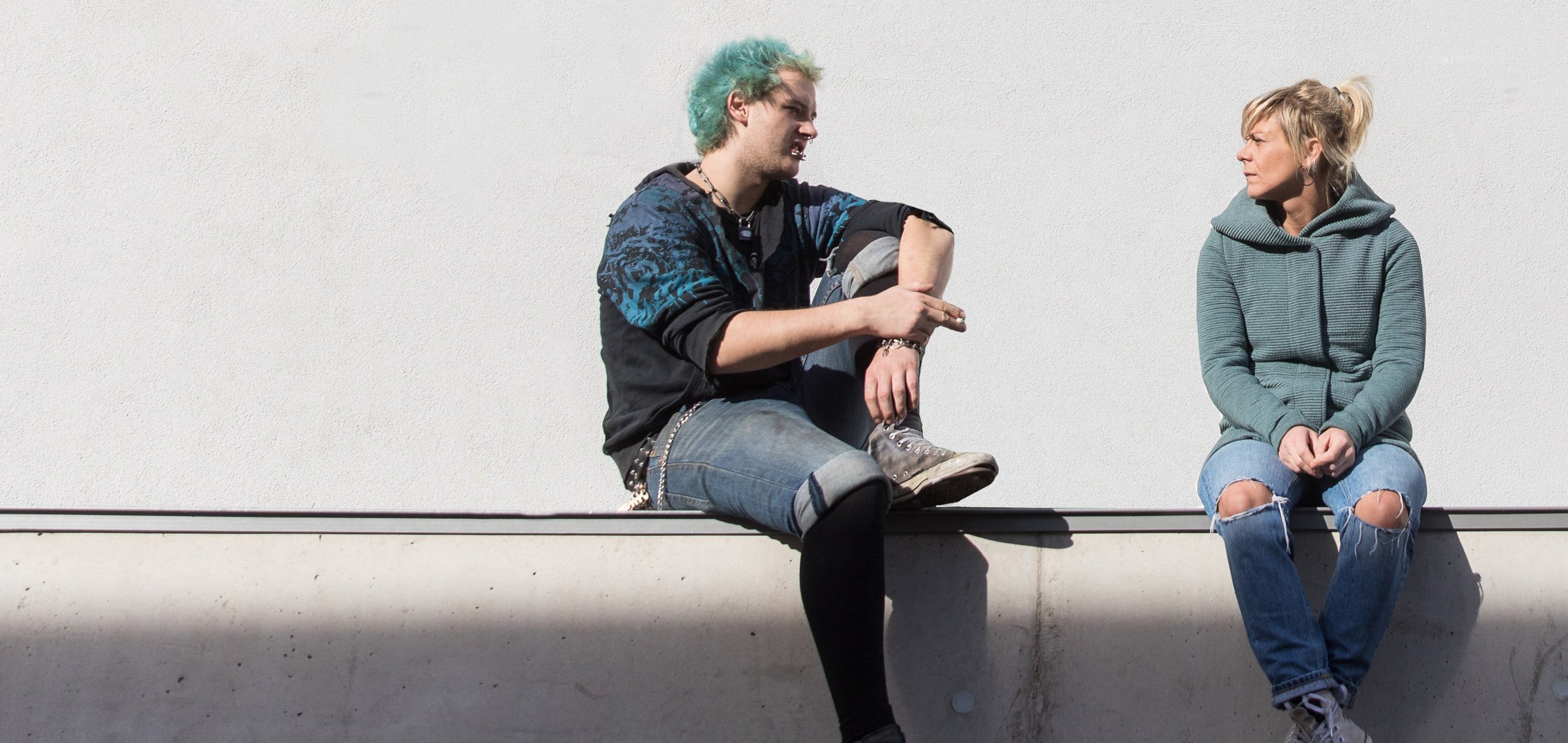 Ein Mann mit tealfarbenen Haaren, eine Zigarette haltend, und eine Frau in grüner Jacke sitzen auf einem Sims vor weißer Wand.