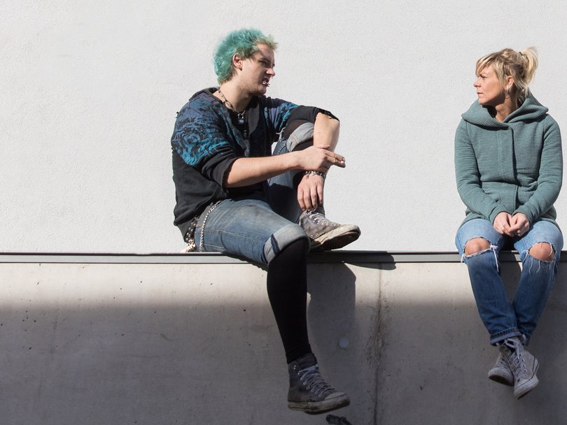 Ein Mann mit tealfarbenen Haaren, eine Zigarette haltend, und eine Frau in grüner Jacke sitzen auf einem Sims vor weißer Wand.