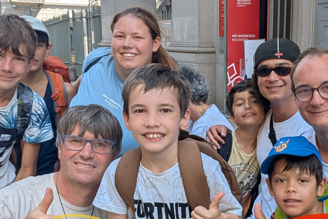 Lachende Gruppe Kinder und Erwachsene vor Gebäude, mit Don Bosco Logo und Text: Sommercamp 2026 in Würzburg.