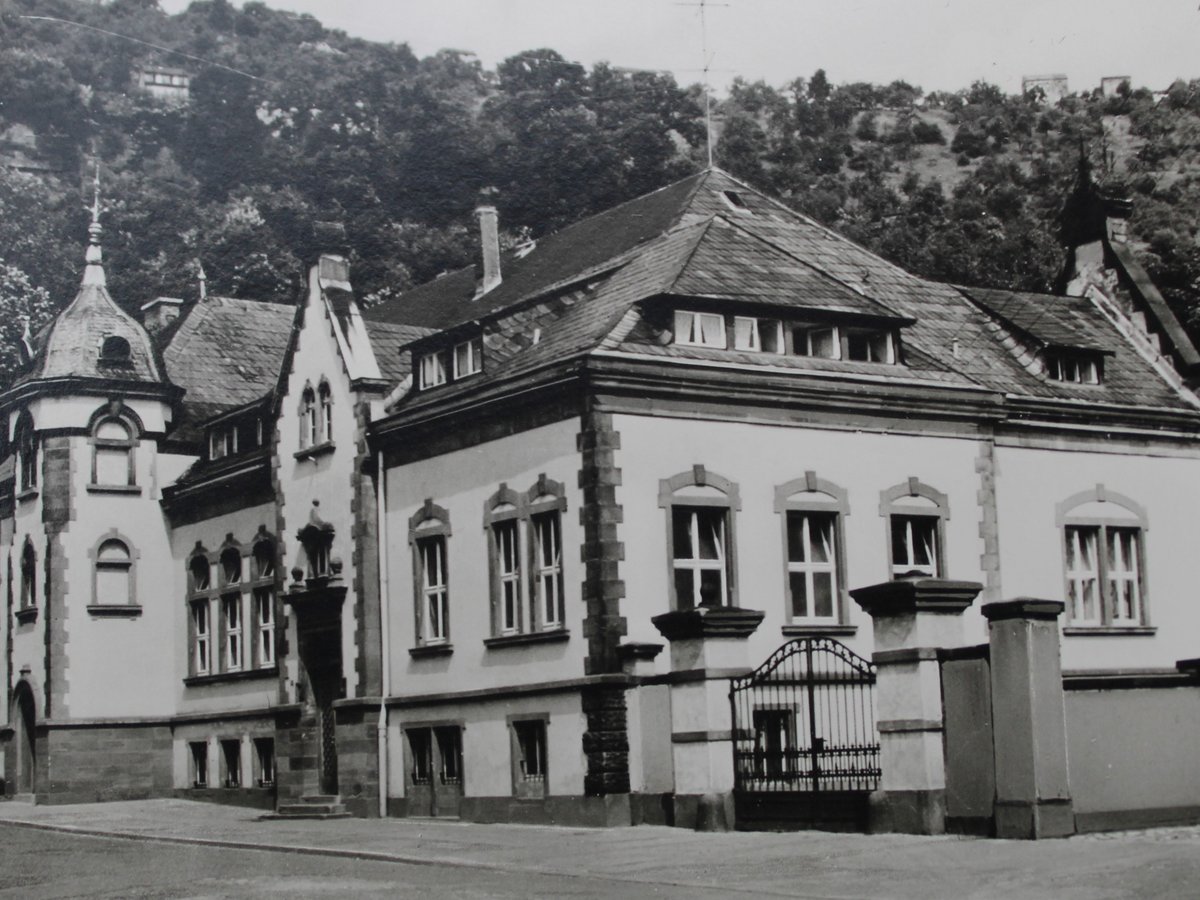 Schwarz-Weiß-Foto einer großen historischen Villa mit Turm und Giebeln an einer Straßenecke, davor ein Oldtimer.