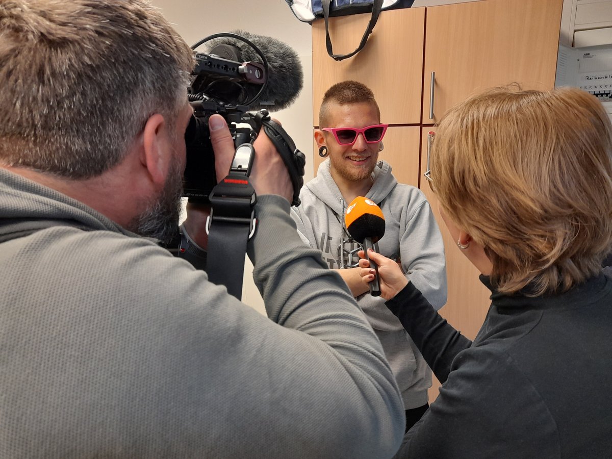 Ein Kameramann filmt einen lächelnden jungen Mann mit pinker Sonnenbrille, der von einer Reporterin mit ZDF-Mikrofon interviewt wird.
