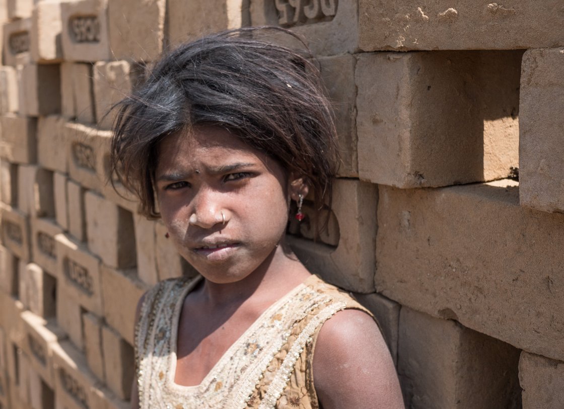 Porträt einer verstaubten jungen Person mit Nasenring, die vor einer Wand aus ungebrannten Ziegeln steht und den Betrachter ansieht.