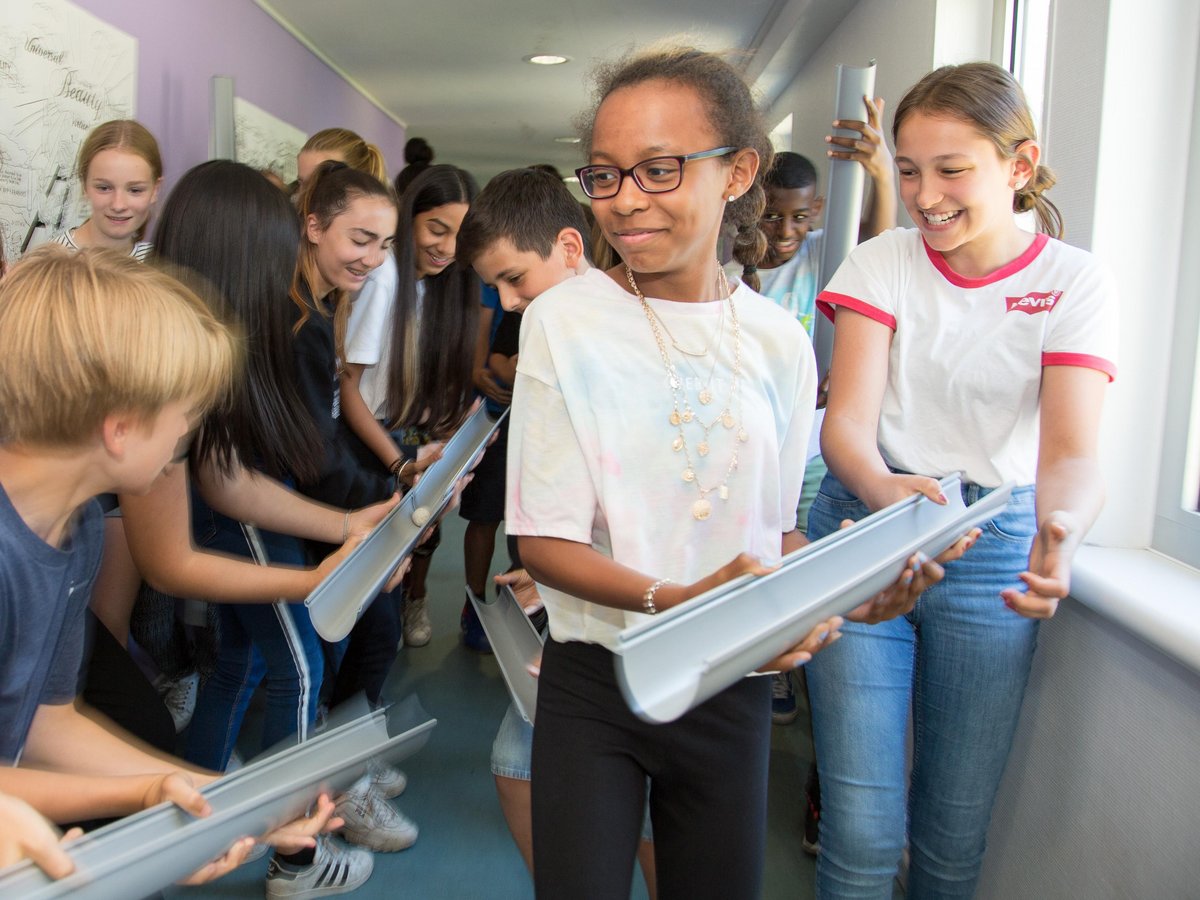 Eine Gruppe verschiedener Schüler lächelt und arbeitet zusammen, um einen Ball durch verbundene Rohre in einer Schule zu führen.