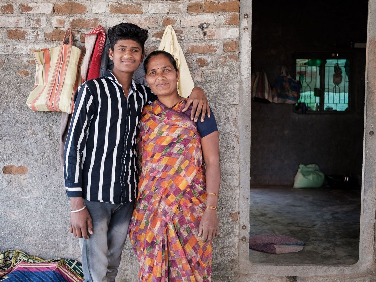 Ein lächelnder junger Mann und eine Frau, wahrscheinlich Mutter und Sohn, posieren Arm in Arm vor einem Haus mit strukturierter Wand.