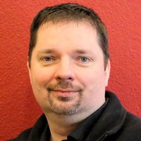 Mann mit dunklen Haaren und Goatee, trägt eine schwarze BVB-Jacke, lächelt vor einer roten Wand.