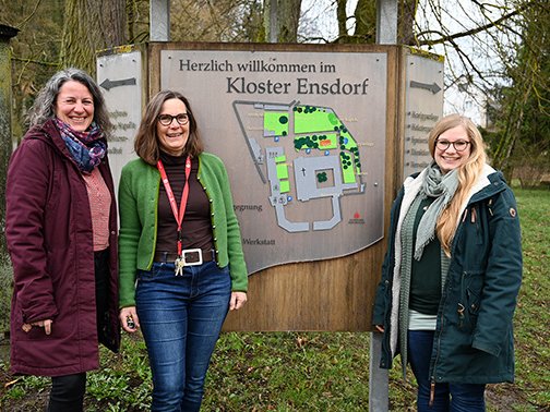Drei lächelnde Frauen stehen vor einem Willkommensschild mit einer Karte des Kloster Ensdorf.