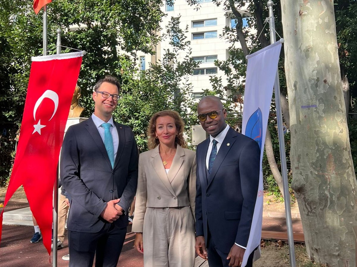 Drei Personen (zwei Männer, eine Frau) lächeln draußen zwischen einer türkischen Flagge und einer anderen Flagge.