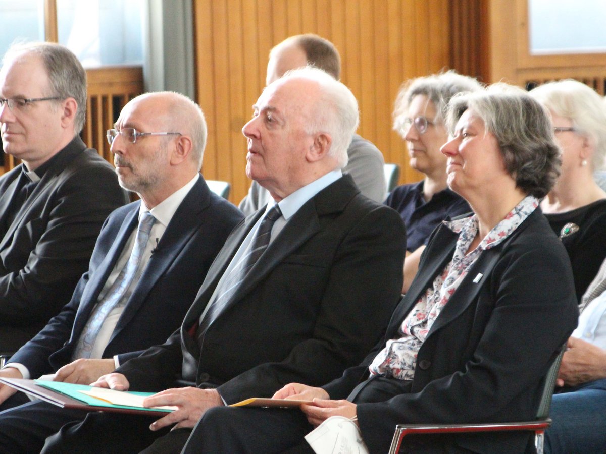 Eine Gruppe von Erwachsenen, Männer und Frauen, sitzen in einer Reihe in einem Innenraum. Einige halten Papiere oder Notizen.