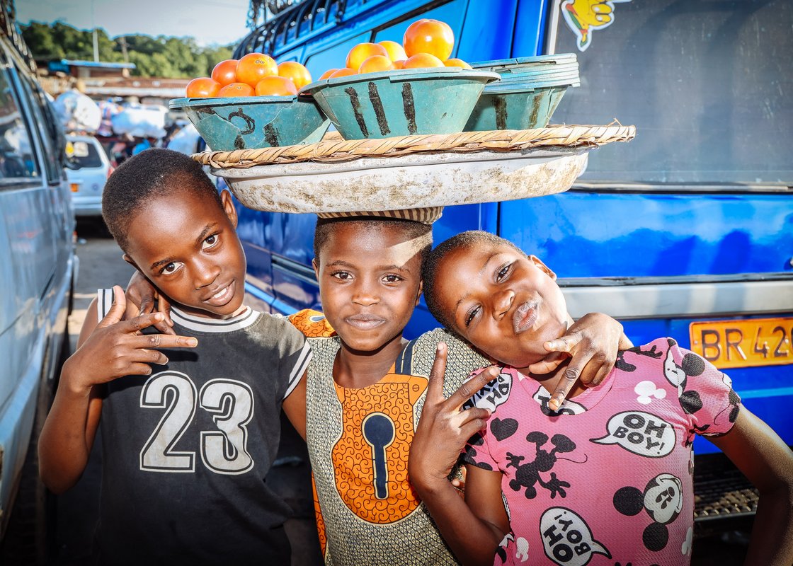 Drei lächelnde Kinder, eines balanciert einen Tomatenkorb auf dem Kopf, vor einem blauen Bus.