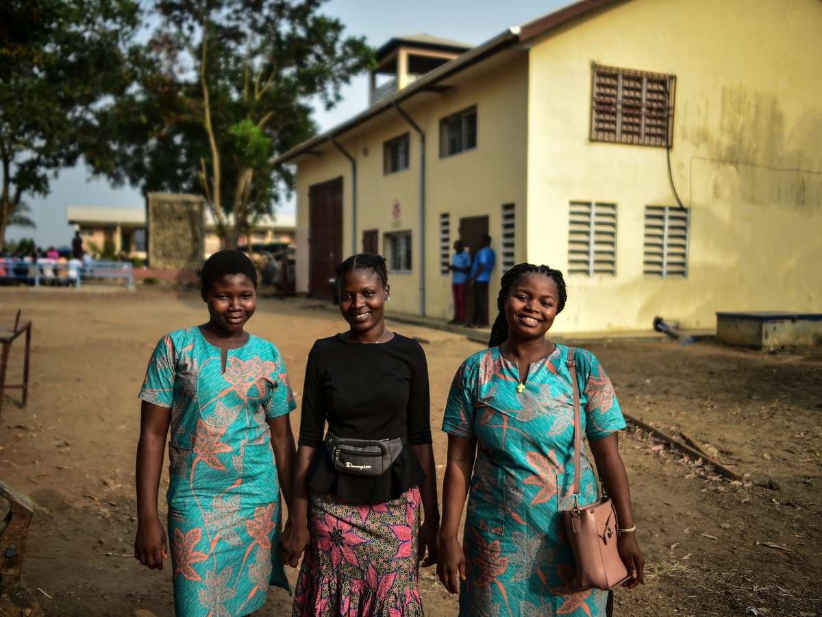Drei lächelnde junge Frauen stehen draußen vor einem gelben Gebäude; zwei tragen passende türkise Kleider.