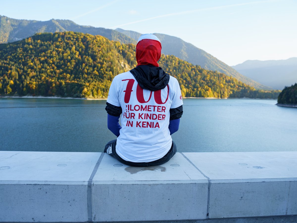 Person von hinten auf einer Betonmauer sitzend, mit T-Shirt '700 Kilometer für Kinder in Kenia', Blick auf See und Herbstberge.