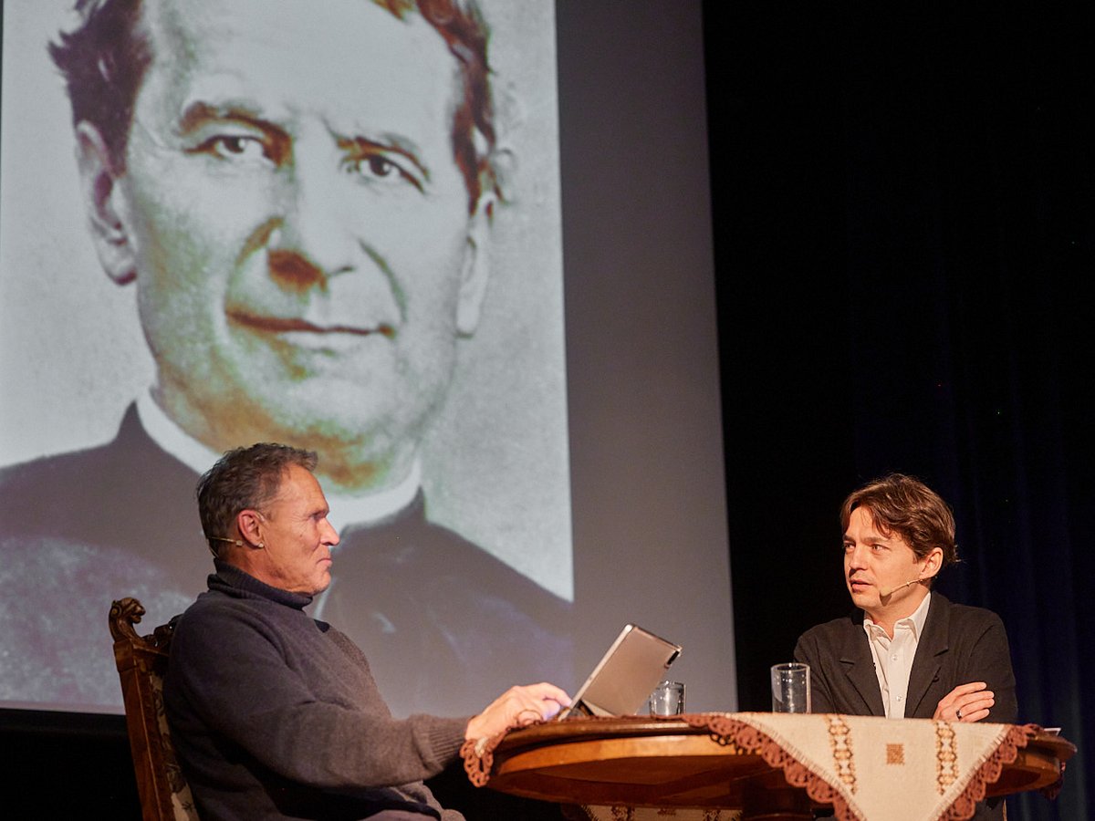 Zwei Männer an einem Diskussionstisch auf einer Bühne, dahinter ein großes Schwarz-Weiß-Porträt eines Priesters.