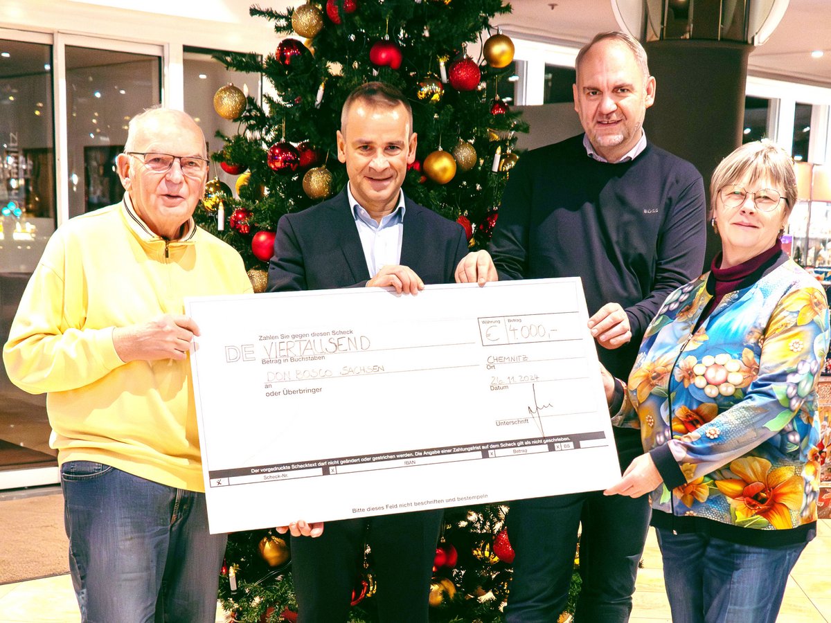 Drei Männer und eine Frau halten einen großen Scheck über 4.000 € für Don Bosco Sachsen vor einem Weihnachtsbaum.