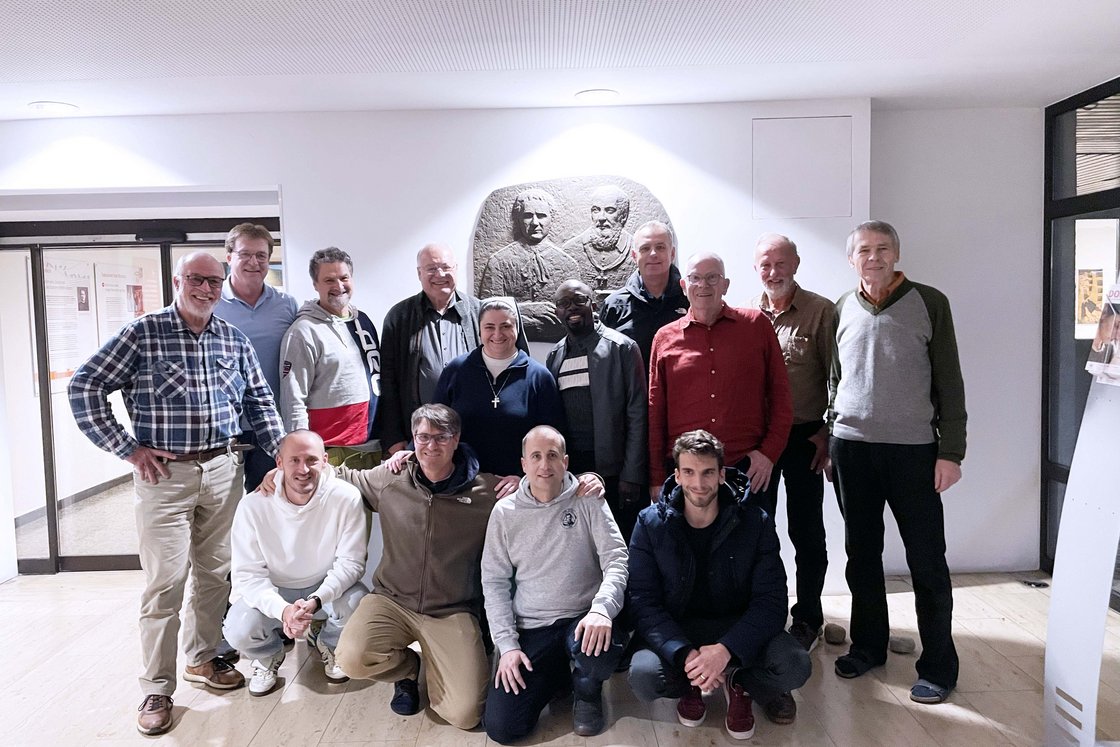 Eine Gruppe von Männern und einer Nonne lächelt für ein Gruppenfoto vor einer großen Wandskulptur mit drei Büsten.