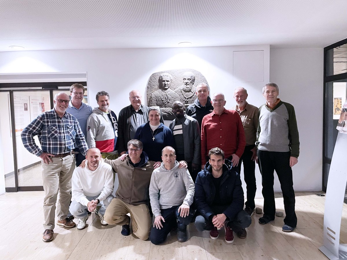 Eine Gruppe von Männern und einer Nonne lächelt für ein Gruppenfoto vor einer großen Wandskulptur mit drei Büsten.